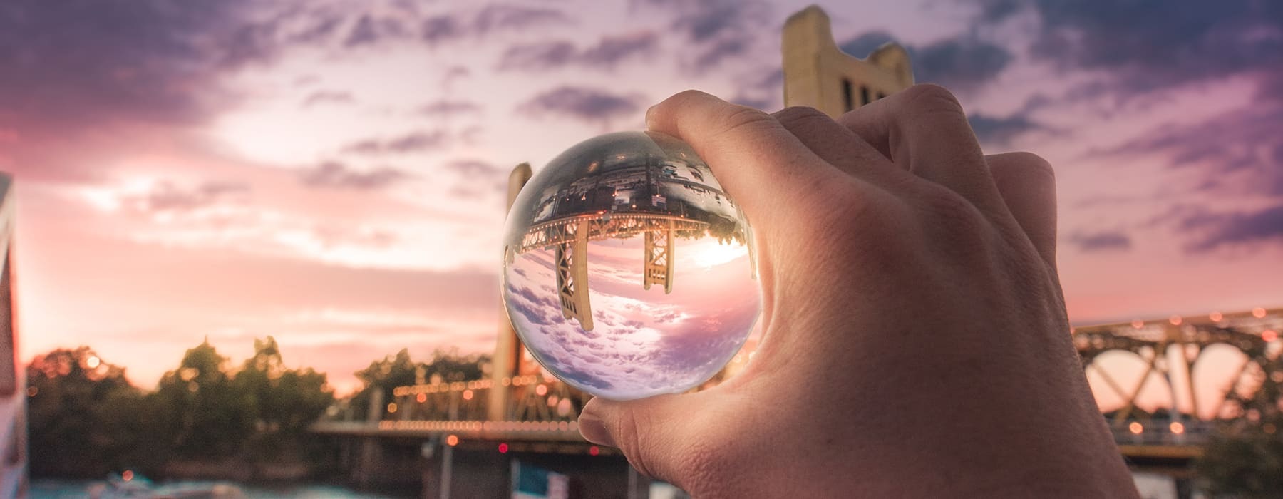 lifestyle image of a hand holding a reflective crystal ball in front of a large river and bridge